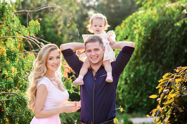 Junge glückliche Familie mit dem kleinen schönen Baby mit den blauen Augen, die im Sommerpark bei Sonnenuntergang gehen