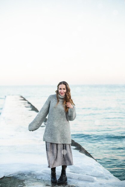 Junge glückliche Frau, die einen Wollpullover trägt, steht im Winter am Strand und genießt den Schneefall