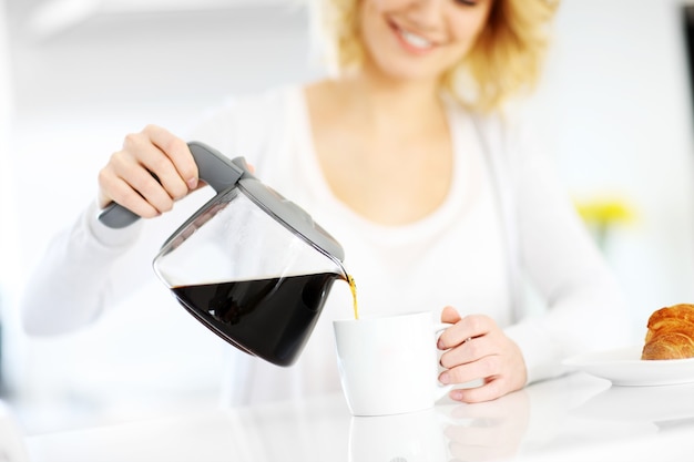 junge glückliche Frau, die in der Küche Kaffee in eine weiße Tasse gießt