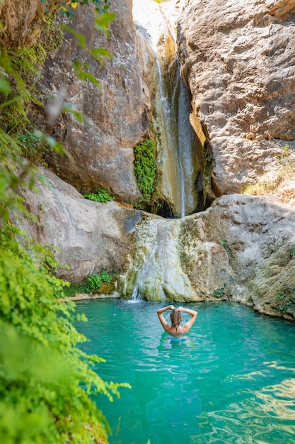 Junge glückliche Frau, die in türkisblauem, kristallklarem Wasser im Fluss mit einem Wasserfall schwimmt, Sommerferien, die Körper und Geist entspannen