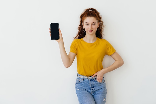 Junge glückliche Frau, die mit dem Finger auf den Bildschirm des Mobiltelefons zeigt und erfreut lächelt, empfiehlt die nützliche App der Anwendung auf weißem Hintergrund