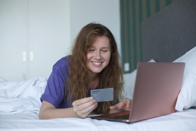 Foto junge glückliche frau, die mit tablet, laptop oder smartphone online einkauft, lag im bett im schlafzimmer