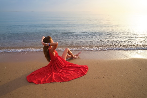 Junge glückliche Frau, die rotes Kleid trägt, das auf Seestrand stillsteht und warmen Sommermorgen genießt.