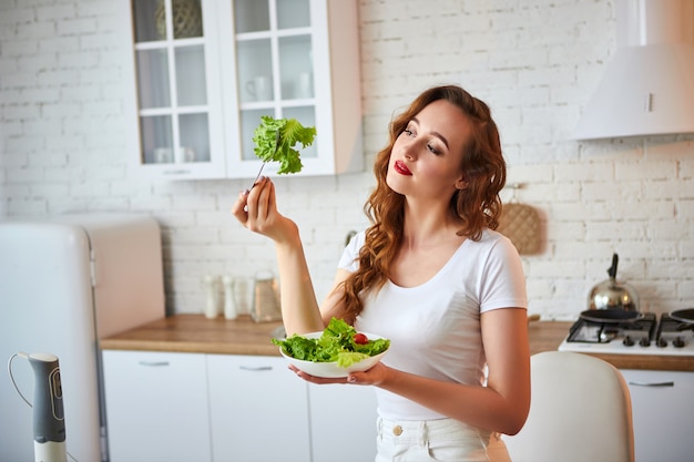 Junge glückliche Frau, die Salat in der schönen Küche mit grünen frischen Zutaten drinnen isst. Gesundes Lebensmittelkonzept