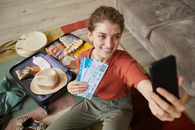 Junge glückliche Frau, die selfie auf ihrem Handy macht, das mit Flugtickets auf dem Boden sitzend aufwirft