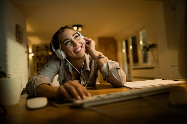 Junge glückliche frau, die spaß hat, während sie computer benutzt und abends zu hause musik über kopfhörer hört