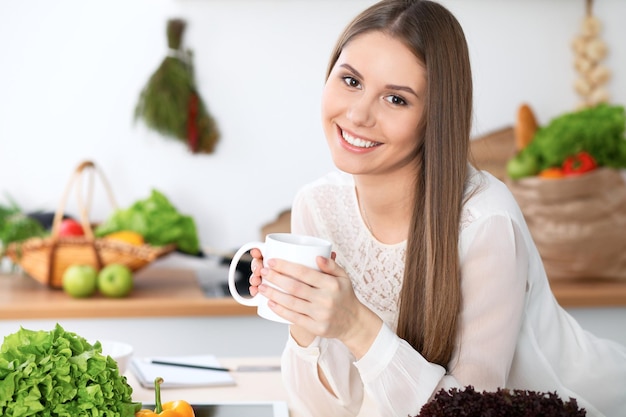 Junge glückliche Frau hält weiße Tasse und schaut in die Kamera, während sie am Holztisch in der Küche zwischen grünem Gemüse sitzt. Hausfrau sucht nach einem neuen Rezept zum Kochen oder Frühstücken