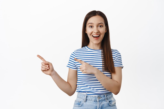 Junge glückliche Frau im T-Shirt, die mit dem Finger nach links auf das Logo zeigt, Verkaufsrabatte zeigt und erstaunt lächelt, Ort oder Veranstaltung empfiehlt, Werbung macht und vor weißem Hintergrund steht.