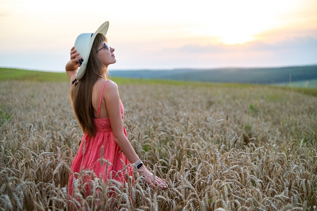 Junge glückliche Frau in rotem Sommerkleid und weißem Strohhut, die auf gelbem Bauernhoffeld mit reifem goldenem Weizen steht und warmen Abend genießt.