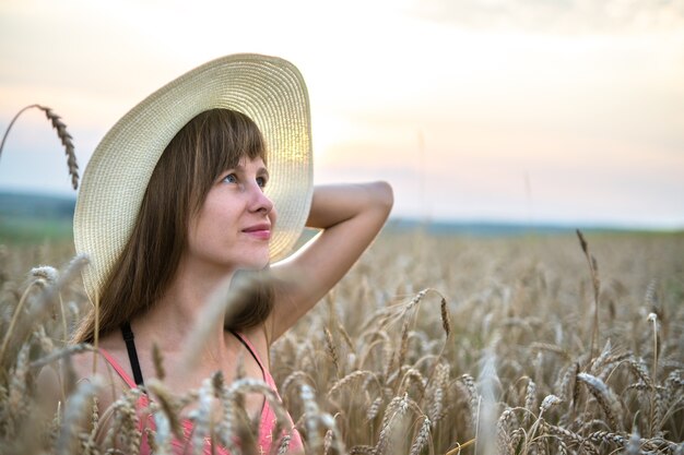 Junge glückliche Frau in rotem Sommerkleid und weißem Strohhut, die auf gelbem Bauernhoffeld mit reifem goldenem Weizen steht und warmen Abend genießt.