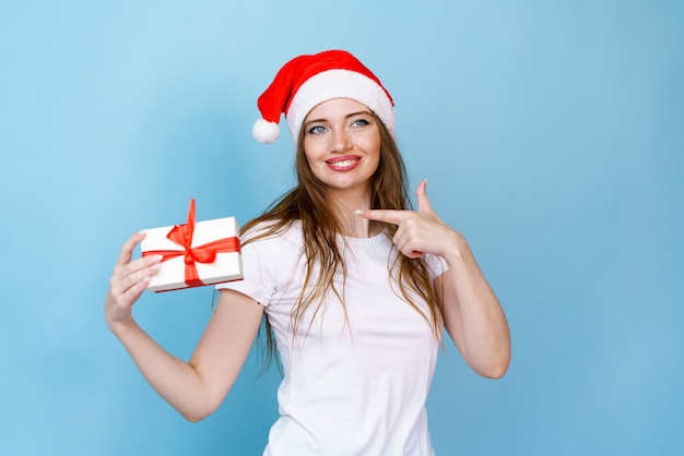 Junge glückliche frau in weihnachtsmütze, die zur seite schaut und weihnachtsgeschenk im weißen t-shirt zeigt und lächelt...