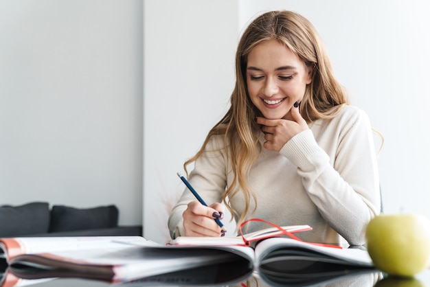 junge glückliche Frau lächelt und macht sich Notizen im Schulheft, während sie am Tisch sitzt
