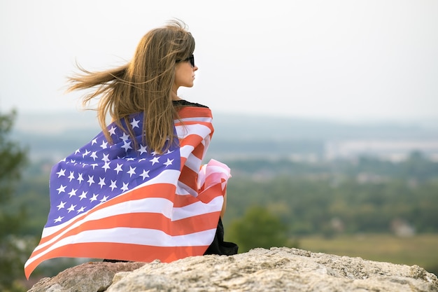 Junge glückliche Frau mit dem langen Haar, das das Winken auf der amerikanischen Windnationalflagge auf ihren Schultern hält, die draußen ruhen, die warmen Sommertag genießen