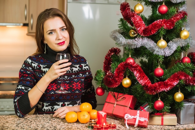 Junge glückliche Frau mit Glas Wein nah an Weihnachtsbaum