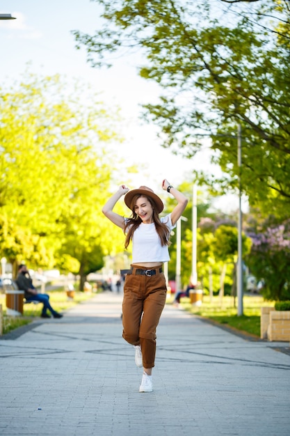 Junge glückliche Frau mit Hut geht durch eine Gasse in einem Park. Ein Mädchen von europäischem Aussehen mit einem Lächeln im Gesicht an einem strahlend sonnigen Sommertag