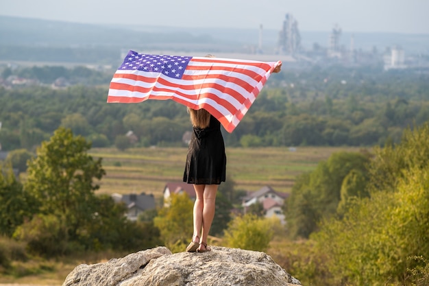 Junge glückliche Frau mit langen Haaren, die auf dem Wind wehende amerikanische Nationalflagge in ihren Händen, die auf einem hohen felsigen Hügel steht und einen warmen Sommertag genießt.