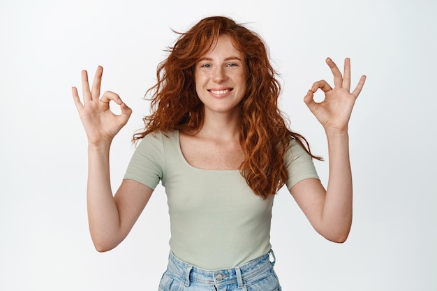 Foto junge glückliche frau mit roten langen haaren zeigt okay-ok-zeichen und sieht erfreut aus, nickt zustimmend und macht eine ja-geste, die im t-shirt auf weißem hintergrund steht