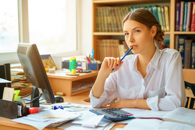 Junge glückliche Geschäftsdame im weißen Hemd, das bei Tisch mit Computer- und Papierarbeitsumgebung sitzt.