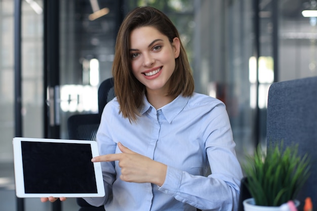 Junge glückliche Geschäftsfrau, die leeren Tablet-Computerbildschirm im Büro zeigt.