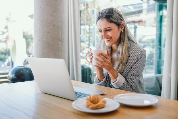Junge glückliche Geschäftsfrau mit Laptop, trinkt Tee oder Kaffee aus einer Tasse an einem modernen offenen Arbeitsplatz