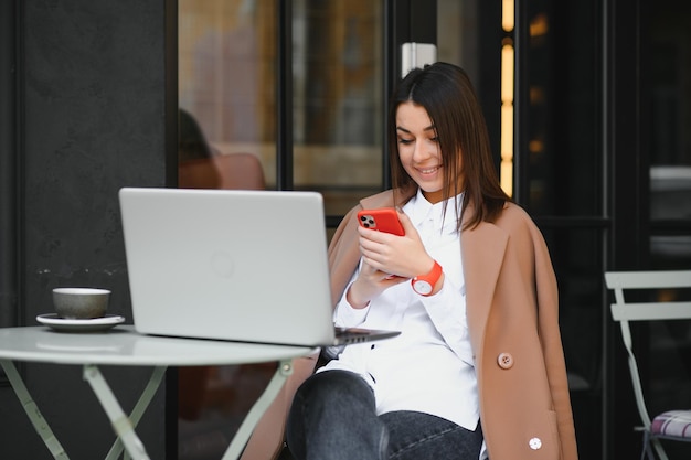 Junge glückliche Geschäftsfrau schaut mit zahnigem Lächeln auf den Smartphone-Bildschirm, während sie mit Laptop und Kaffee draußen auf der Terrasse sitzt, aus der Ferne online Berichte überprüft und sich auf ein Treffen vorbereitet