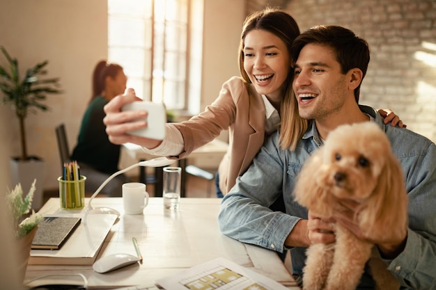 Junge glückliche Kollegen haben Spaß beim Telefonieren und machen Selfies mit einem Hund im Büro