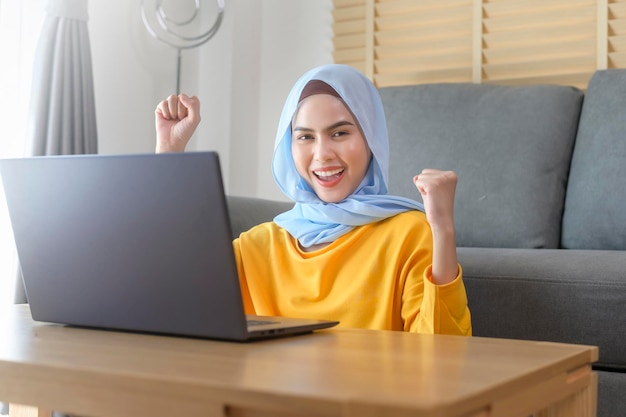 Junge glückliche muslimische Frau, die zu Hause am Laptop im Wohnzimmer arbeitet