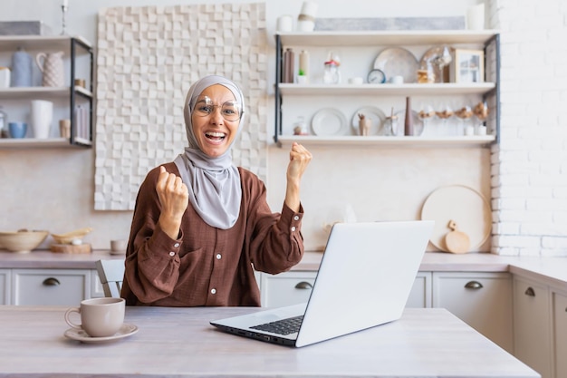 Junge glückliche muslimische studentin im hijab, die zu hause sitzt und auf dem laptop studiert, zeigt eine ja-geste