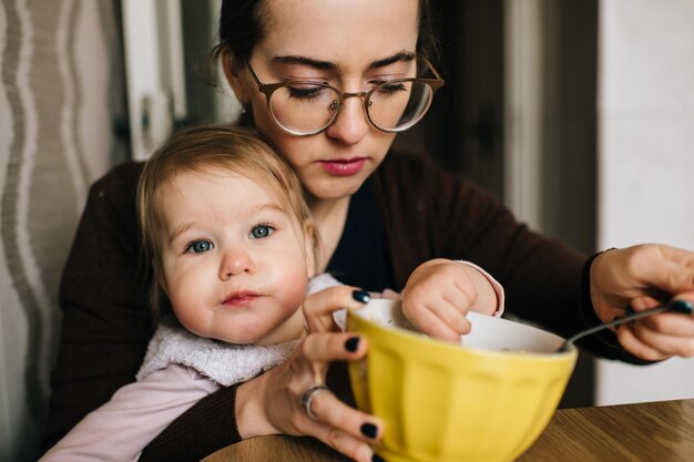 Junge glückliche Mutter, die ihr kleines Baby am Tisch füttert.