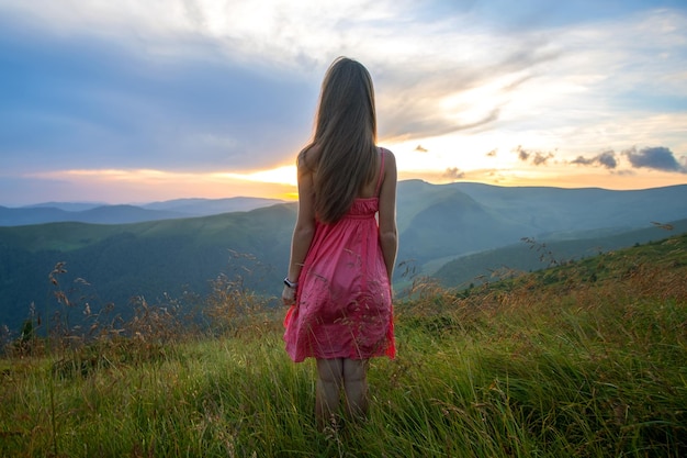 Junge, glückliche Reisende im roten Kleid, die an einem windigen Abend in den Sommerbergen auf einem grasbewachsenen Hang steht und bei Sonnenuntergang den Blick auf die Natur genießt.