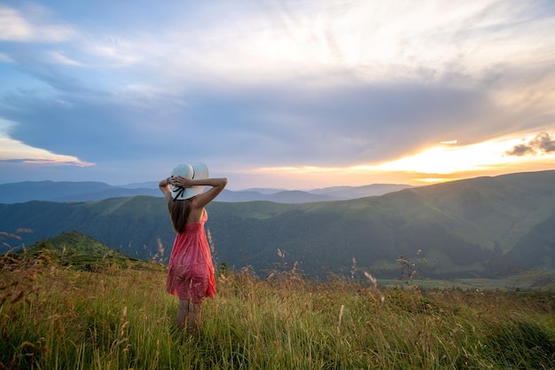 Junge glückliche Reisende in rotem Kleid, die an einem windigen Abend in den Sommerbergen auf einem grasbewachsenen Hügel steht und bei Sonnenuntergang den Blick auf die Natur genießt.