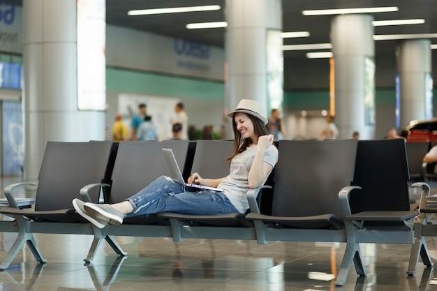 Junge glückliche Reisende Touristenfrau sitzt am Laptop, macht Gewinnergeste und wartet in der Lobbyhalle am internationalen Flughafen?