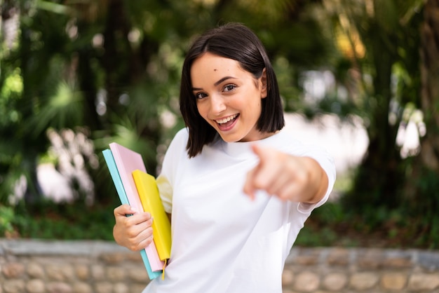 Junge glückliche Studentin, die nach vorne zeigt und Notizbücher im Freien hält