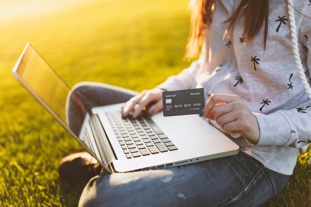 Junge glückliche Studentin mit Kreditkarte. Frau, die auf Grasboden sitzt und an Laptop-PC im Stadtpark auf grünem Grassonnenscheinrasen im Freien arbeitet. Mobiles Büro. Freiberufliches Geschäftskonzept.