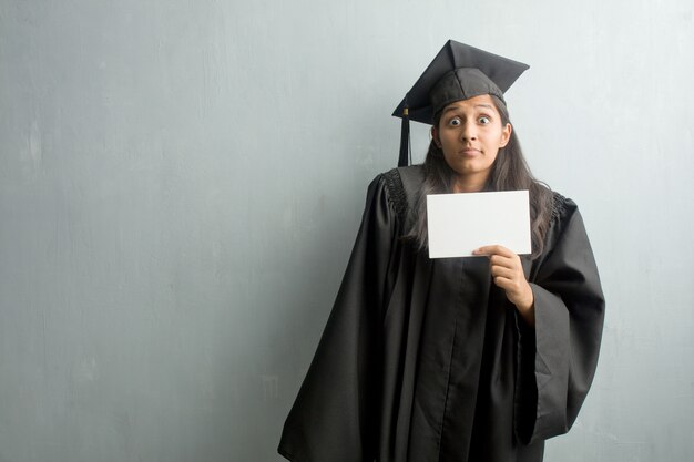 Junge graduierte indische Frau gegen eine Wand, die Schultern zweifelt und zuckt