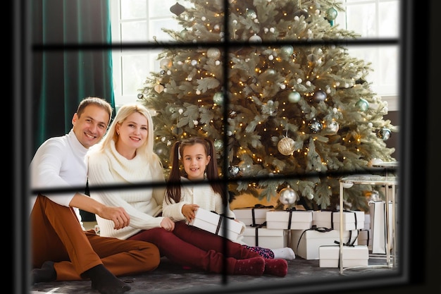 Junge große Familie feiert Weihnachten beim Abendessen, Blick von außen durch ein Fenster in ein geschmücktes Wohnzimmer mit Baum- und Kerzenlichtern, glückliche Eltern essen mit drei Kindern.