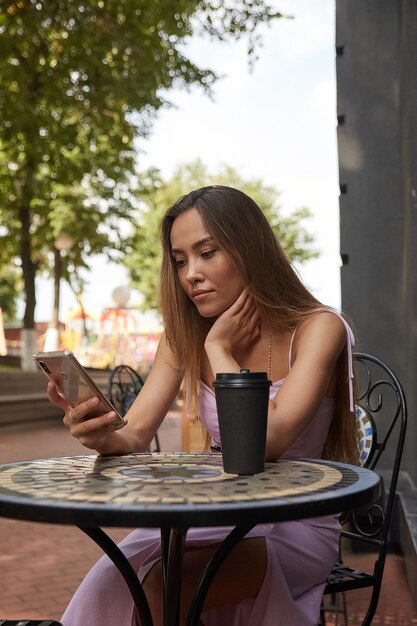 Junge gut gekleidete asiatische Frau sitzt am Tisch mit Kaffee mit Smartphone