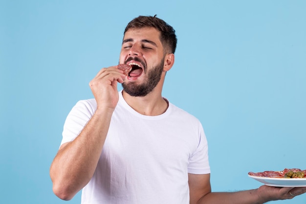 Junge hält einen Teller mit Salami und isst es auf blauem Hintergrund Mann isst Salami