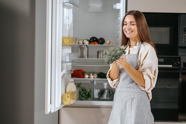 Junge Hausfrau, die nahe dem Kühlschrank in der Küche steht