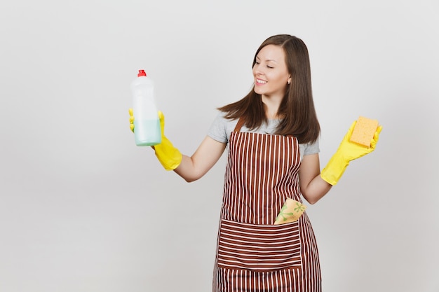 Junge Hausfrau in gelben Handschuhen, gestreifter Schürze, Putzlappen in der Tasche isoliert auf weißem Hintergrund. Frau, die eine Flasche mit sauberer Flüssigkeit zum Abwaschen von Geschirr hält, Schwamm. Kopieren Sie Platz für Werbung.