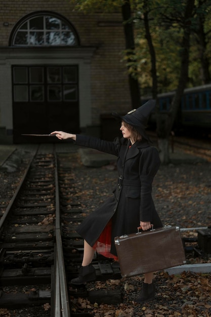 Junge Hexe mit Zauberstab zaubert am alten Bahnhof Mädchen mit schwarzem Umhang und Spitzhut und hält Retro-Koffer in den Händen Vertikaler Rahmen