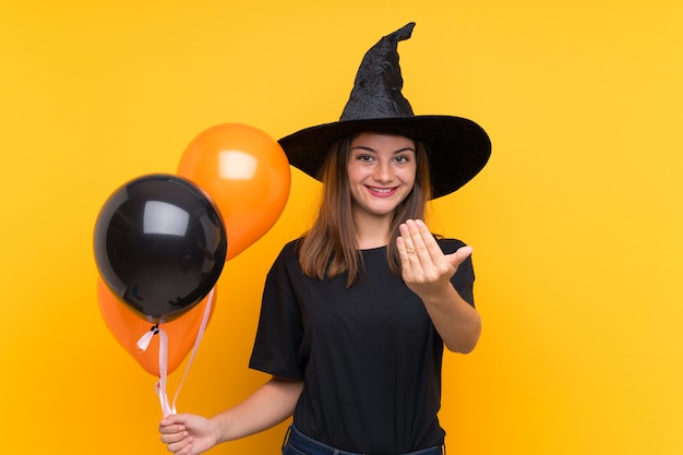 Junge Hexe, welche die schwarzen und orange Luftballone für Halloween-Parteien einlädt, mit der Hand zu kommen hält. Schön, dass Sie gekommen sind