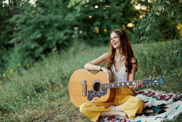 Junge Hippie-Künstlerin spielt Gitarre und singt Lieder in umweltfreundlicher Kleidung, sitzt im Herbst draußen in der Natur auf dem Boden und blickt auf den Sonnenuntergang. Hochwertiges Foto