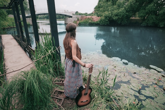 Junge Hippiefrau mit der Gitarre, die nahe dem Teich steht