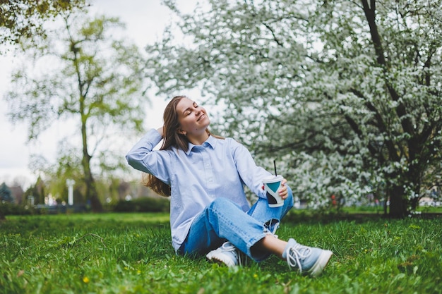 Junge Hipster-Frau in Jeansjacke ruht auf grünem Rasen im Park und trinkt Kaffee, der sich im Freien entspannt Lifestyle-Kaffeepause Selektiver Fokus auf weibliche Hände, die eine Einwegtasse Kaffee halten