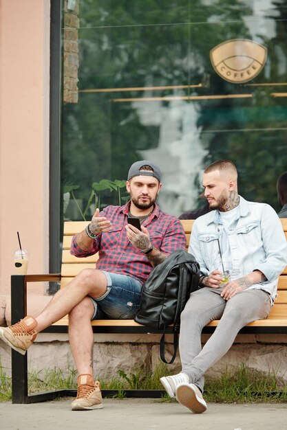 Junge Hipster-Männer sitzen auf Bank nahe Coffeeshop und diskutieren Internetnachrichten