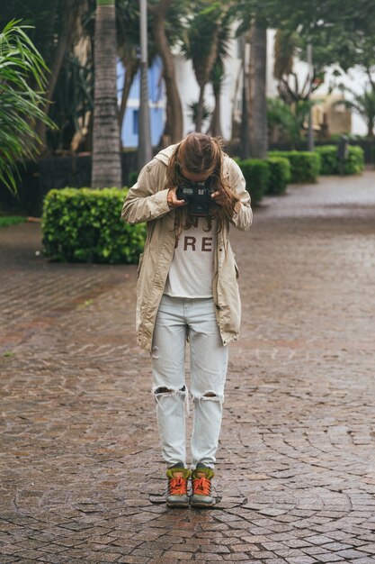 Junge Hipsterfrau, die Reflexkamera hält und Fotos von ihren Stiefeln auf nassem Steinpflaster macht