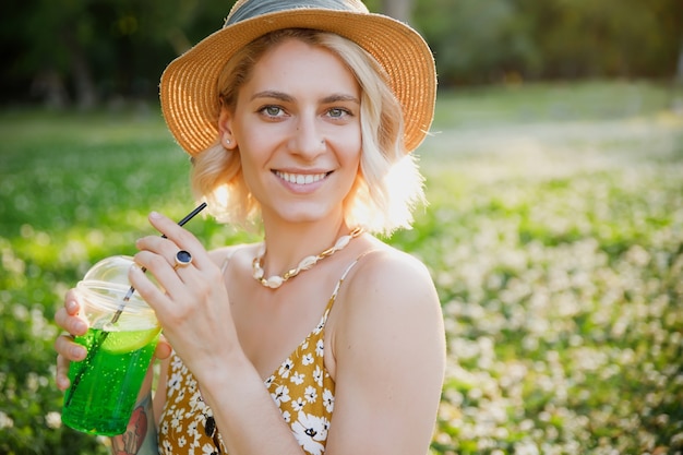 Junge Hipsterfrau im Sommerkleid und im Hut mit Sommeraccessoires und Cocktail