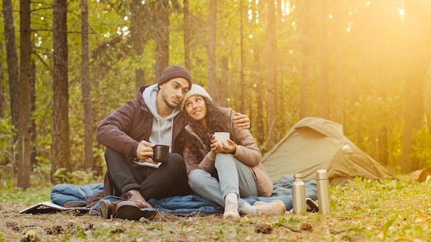 Junge hispanische Ehepaartouristen ruhen sich im Wald aus und campen Mann und Frau in der Liebe, die heißen Tee trinkt