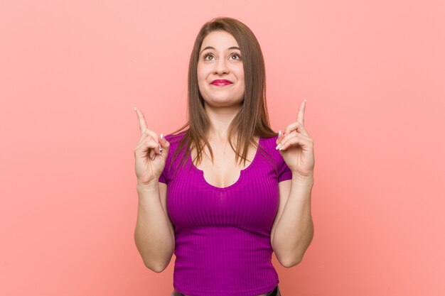 Junge hispanische Frau auf rosa Wand zeigt mit beiden Vorderfingern an, die sich eine Leerstelle zeigen.